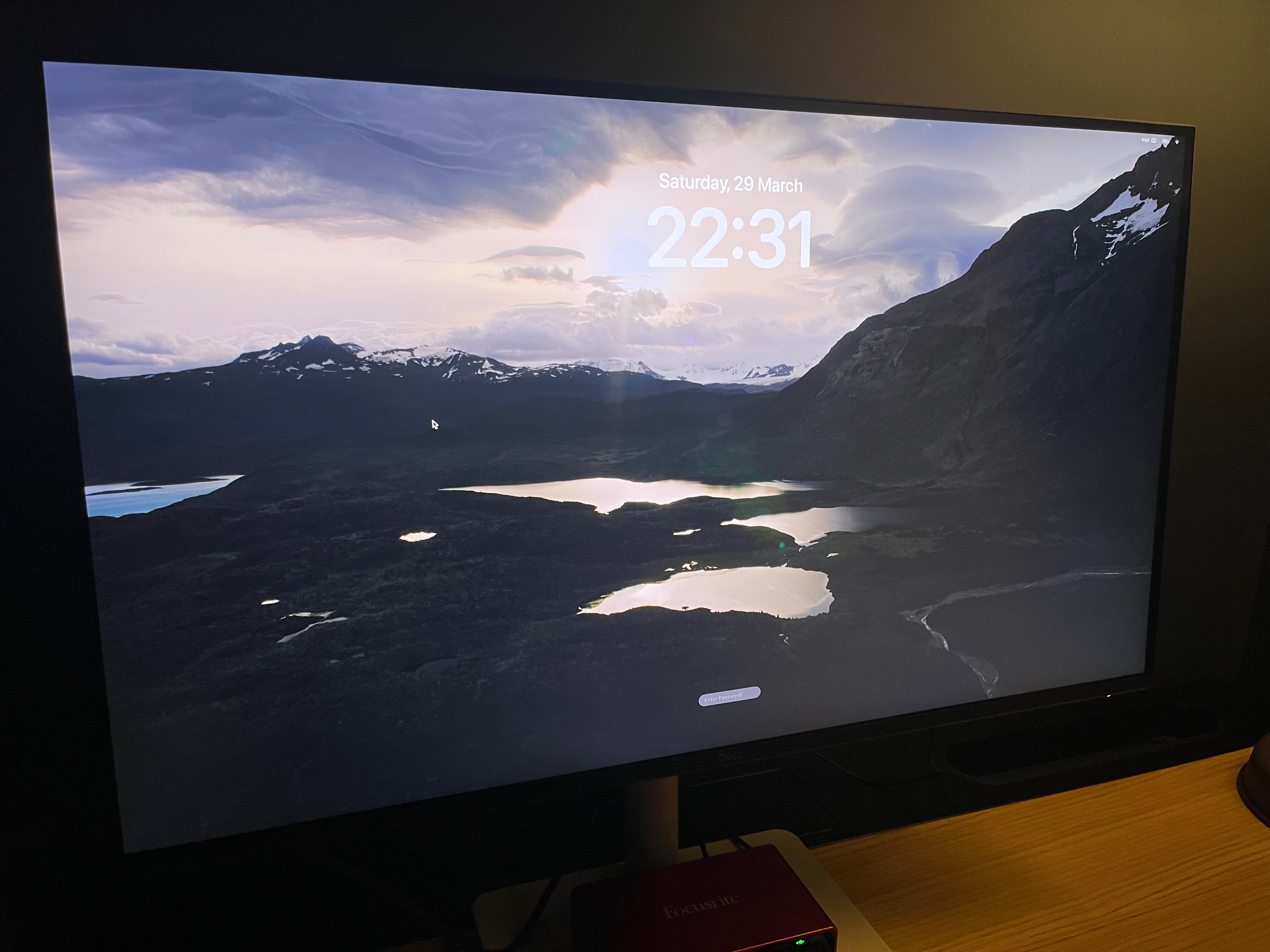 The monitor turned on displaying a macOS screensaver. Sitting below the monitor on its stand is a red Focusrite audio interface with cables connected.